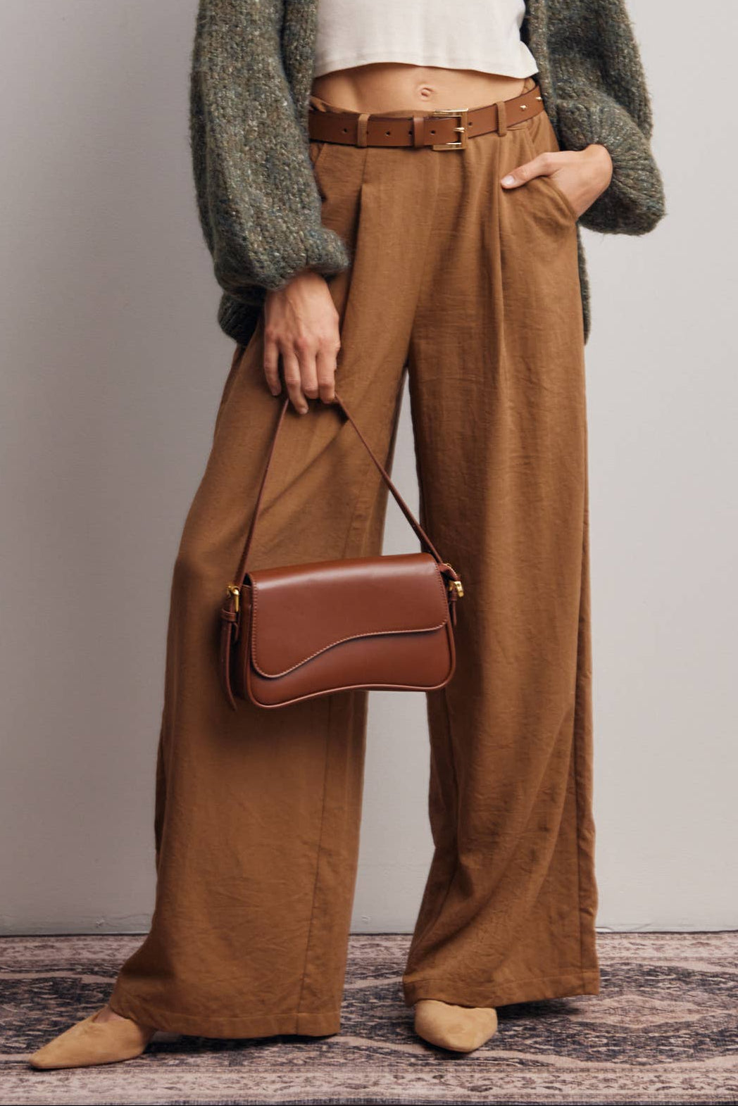 Woman wearing a green cardigan, white shirt, and brown pants holding a brown handbag on a plain background