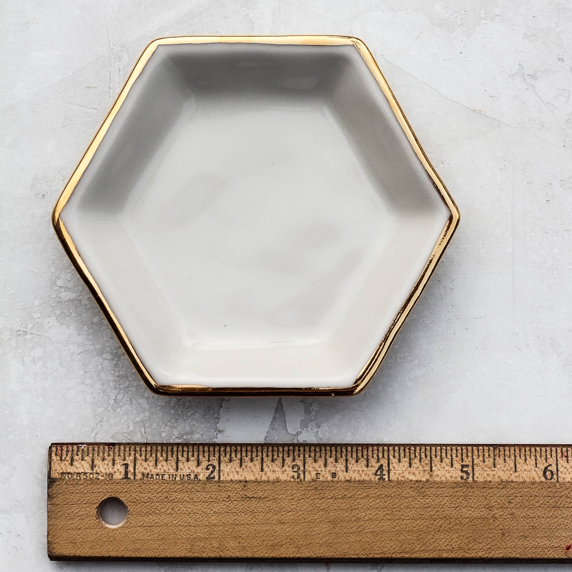 Hexagonal ceramic dish with gold rim on a light gray surface, measured by a wooden ruler.