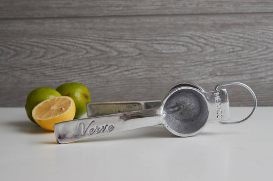 Metal measuring spoon with 'Verve' branding next to limes on a wooden surface
