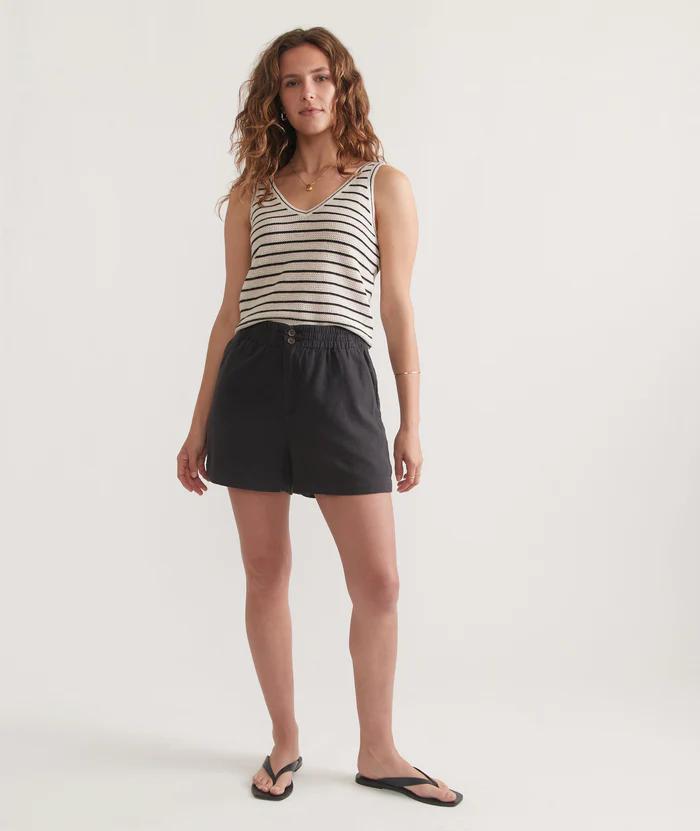 A woman standing against a white background, wearing a striped tank top and faded black shorts.