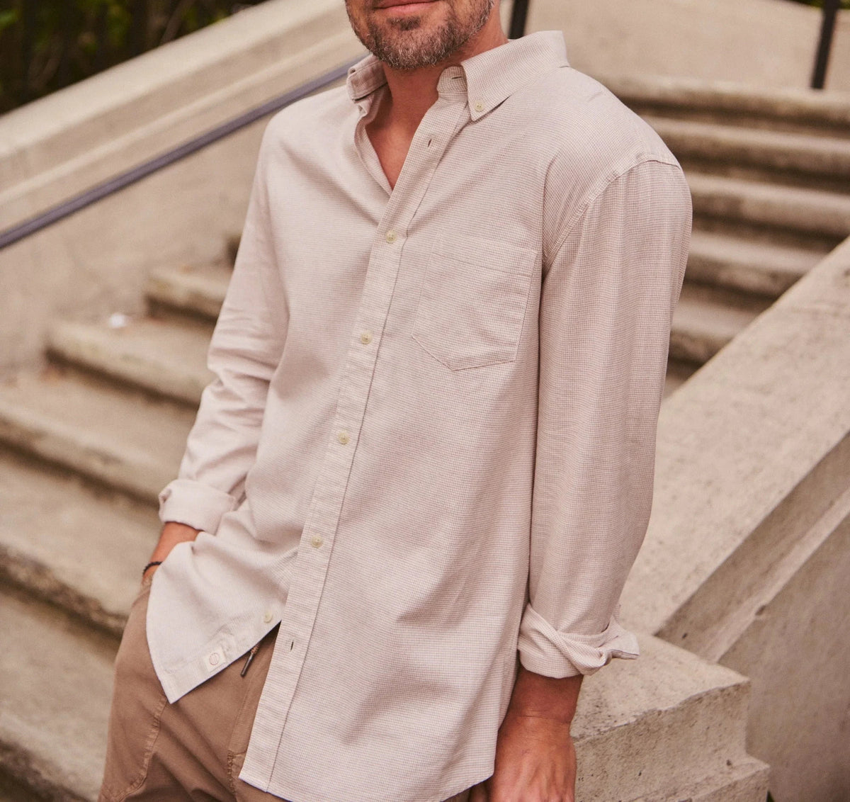 A man wearing a light beige long-sleeve linen button-down shirt and a navy blue baseball cap, standing on outdoor concrete stairs.