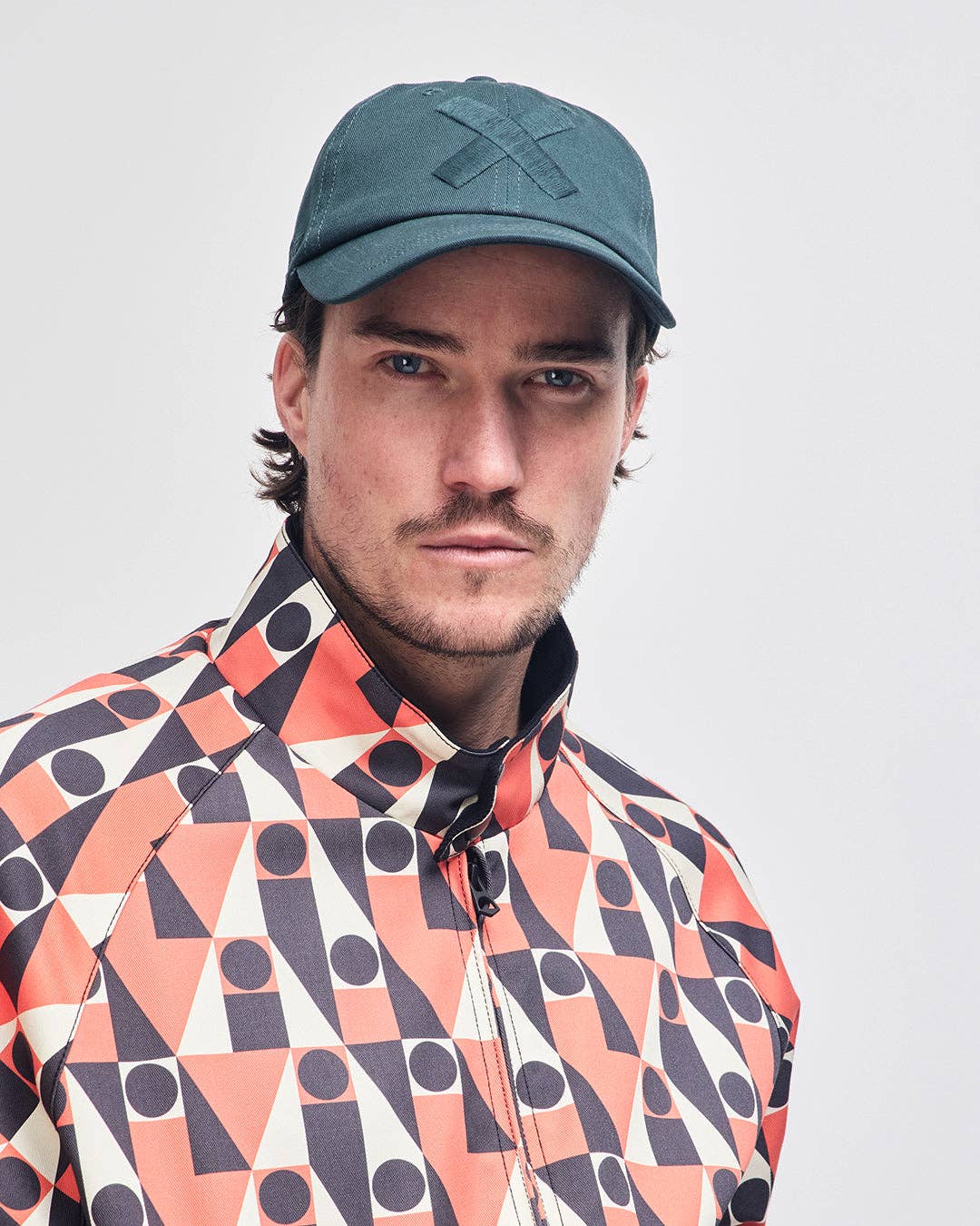 Man wearing a patterned jacket and green cap against a plain background