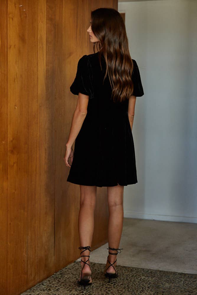 Woman wearing a black dress standing in a room with wooden walls and a white door.