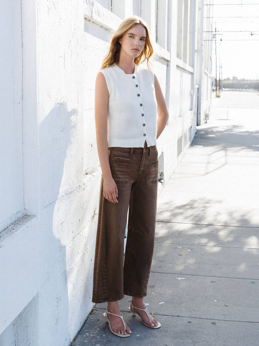 Woman wearing a white sleeveless sweater and brown pants standing against a white wall.