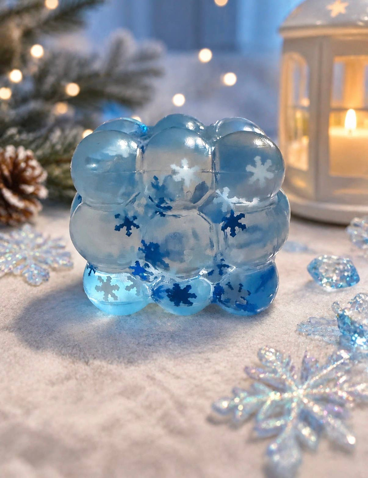 A cluster of translucent light blue and medium blue gel-like spheres with white snowflake patterns, arranged on a snowy surface with holiday decorations, a lit lantern, and blurred lights in the background.