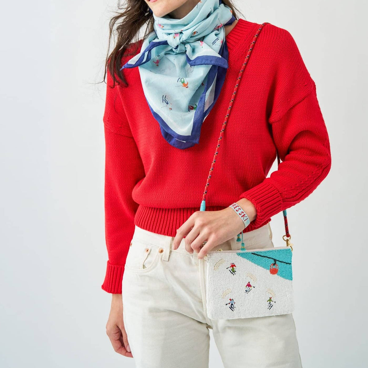 A woman wearing a bright red knit sweater, cream pants, a light blue patterned scarf, and a matching light blue and white crossbody bag with small embroidered details.