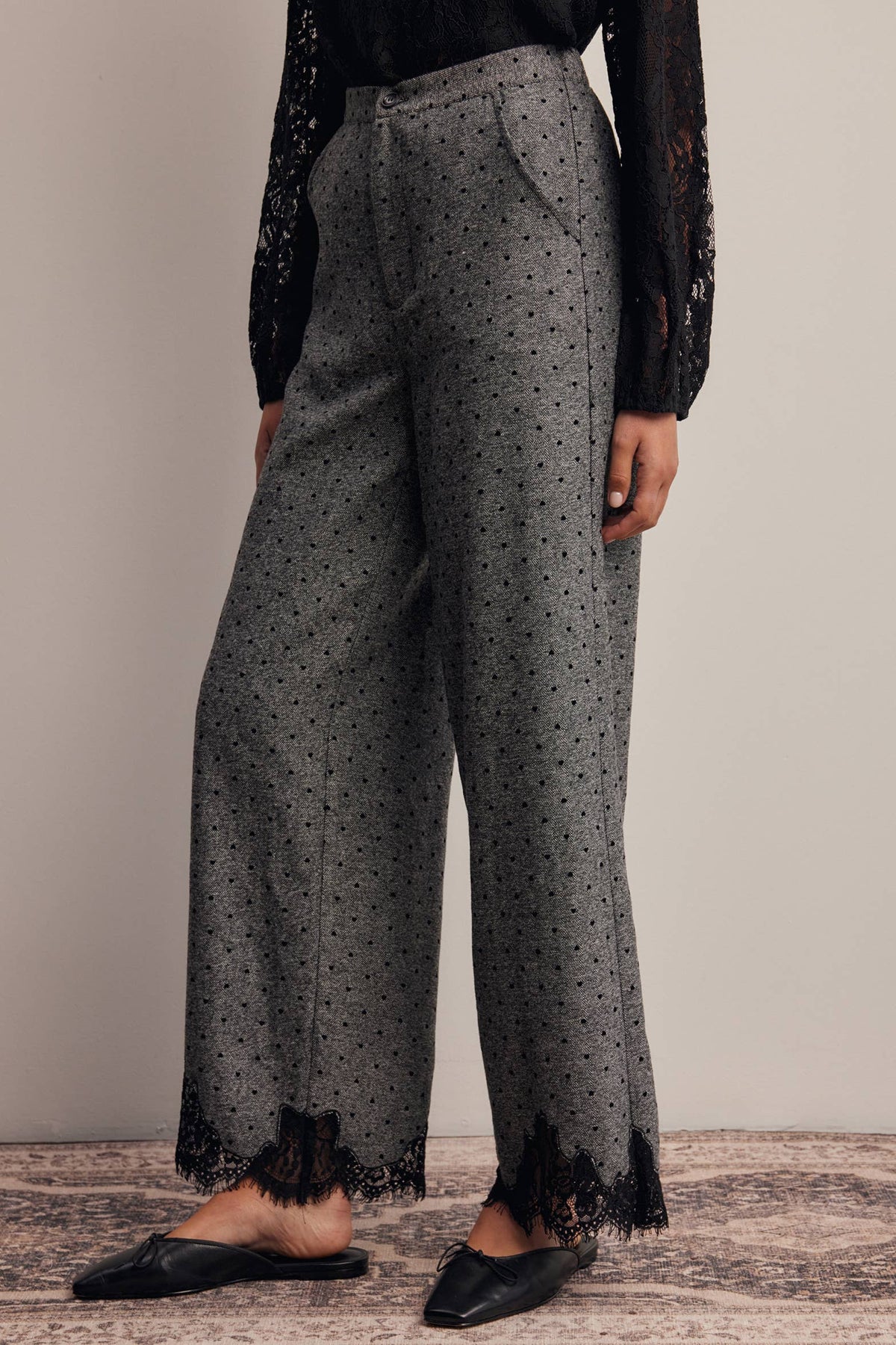 A person wearing high-waisted wide-leg gray pants with a small black dot pattern and black lace trim at the hems, paired with black pointed flats, against a neutral studio backdrop.