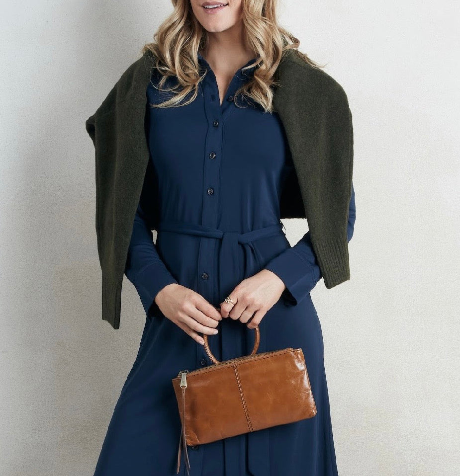 A woman wearing a navy blue long-sleeve button-front dress with a matching waist tie and an olive green shrug draped over her shoulders, holding a brown leather handbag against a light neutral wall.