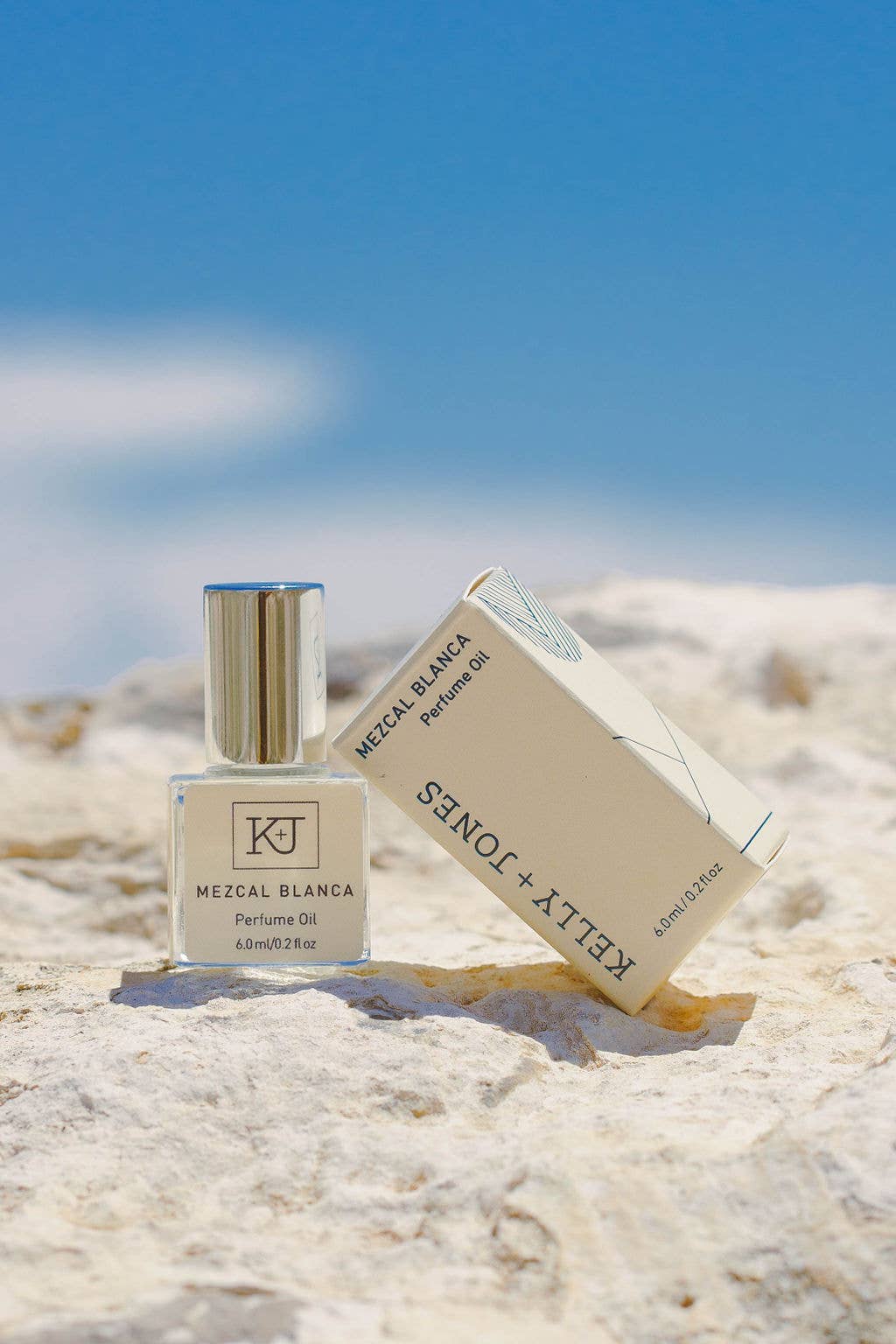 KJ Mezcal Blanca perfume bottle and packaging on a sandy surface with a blue sky background