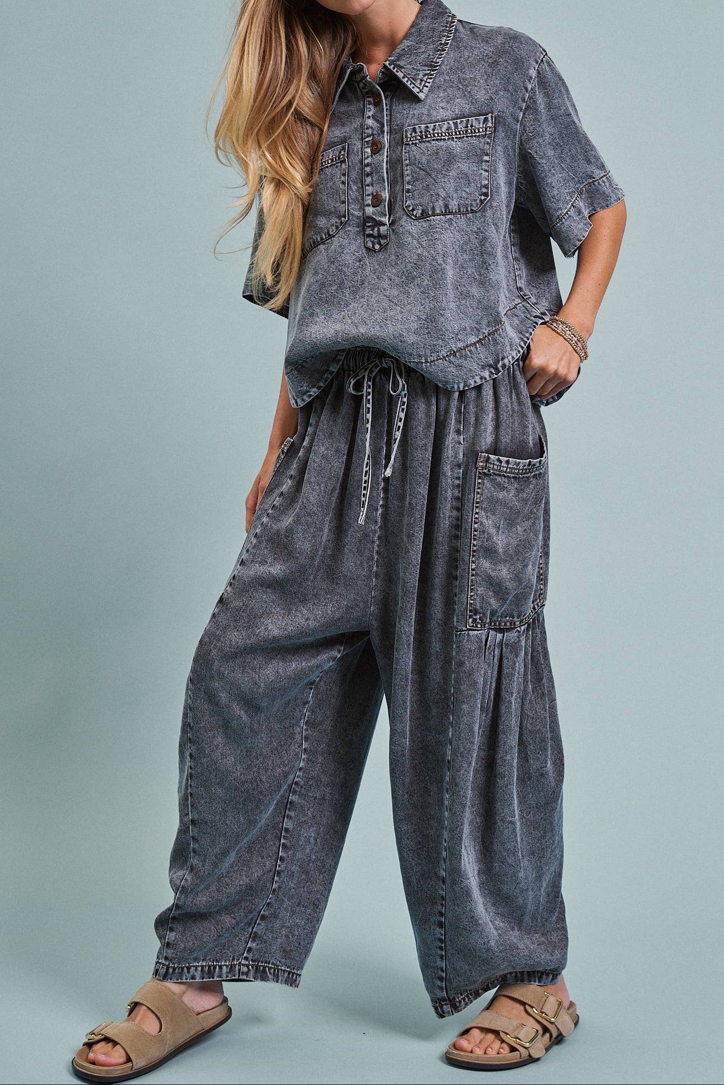 A woman modeling a matching acid-wash denim two-piece set consisting of a short-sleeve button-up shirt and wide-leg pants, paired with tan slide sandals against a solid light blue-gray studio background.