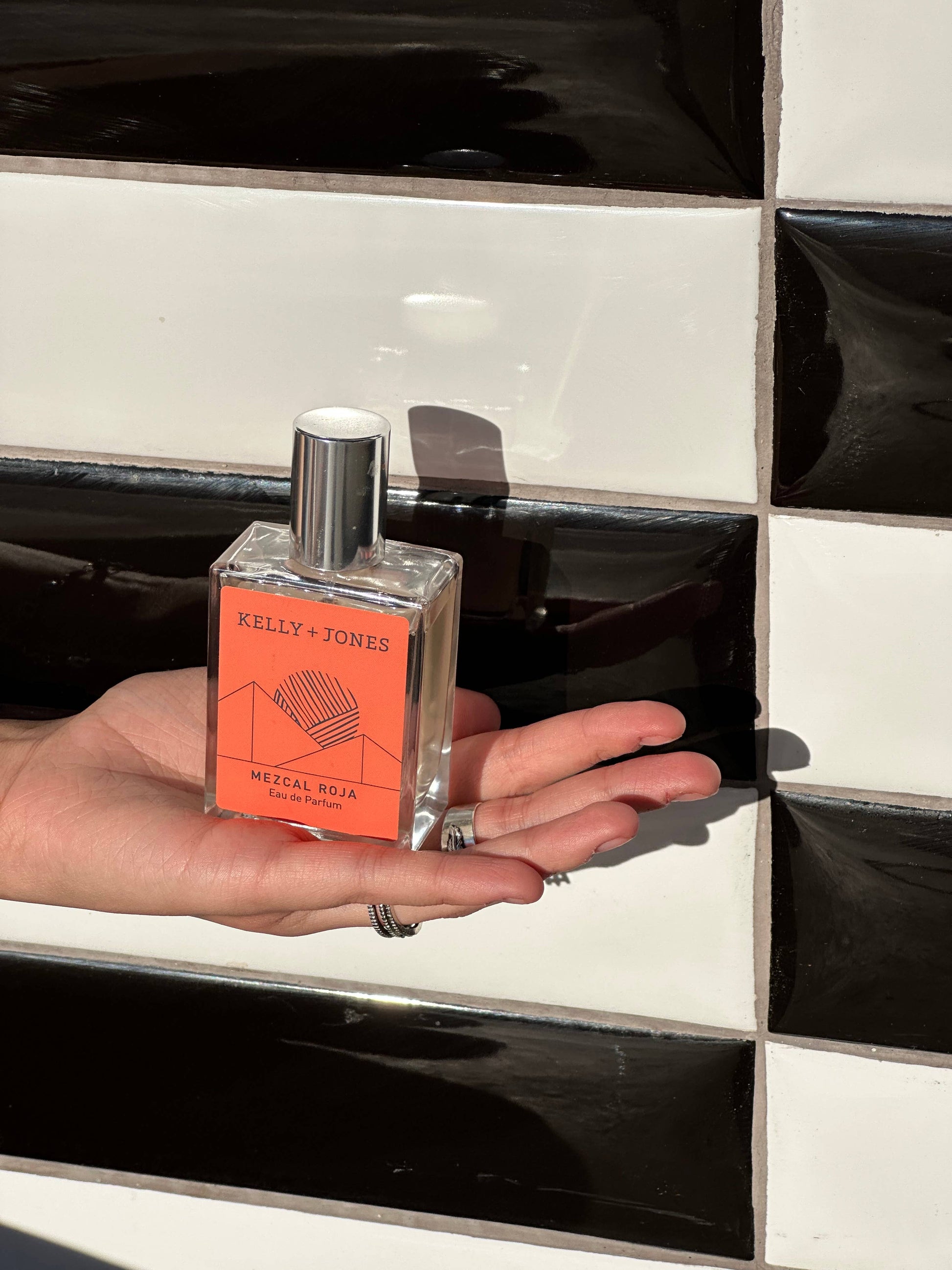 Person holding a Kelly & Jones perfume bottle against a black and white tiled wall.