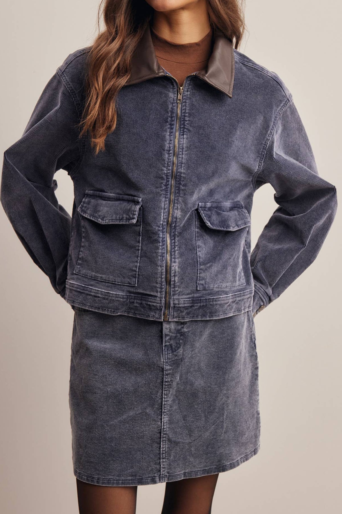 A woman modeling a matching corduroy jacket and skirt set in a washed slate blue color, featuring a zip-up jacket with flap pockets and a matching mini skirt, set against a plain beige studio background.