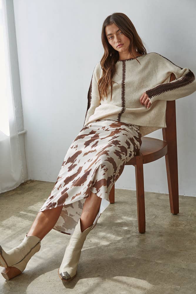 Woman sitting on a wooden chair wearing a beige sweater and patterned skirt.