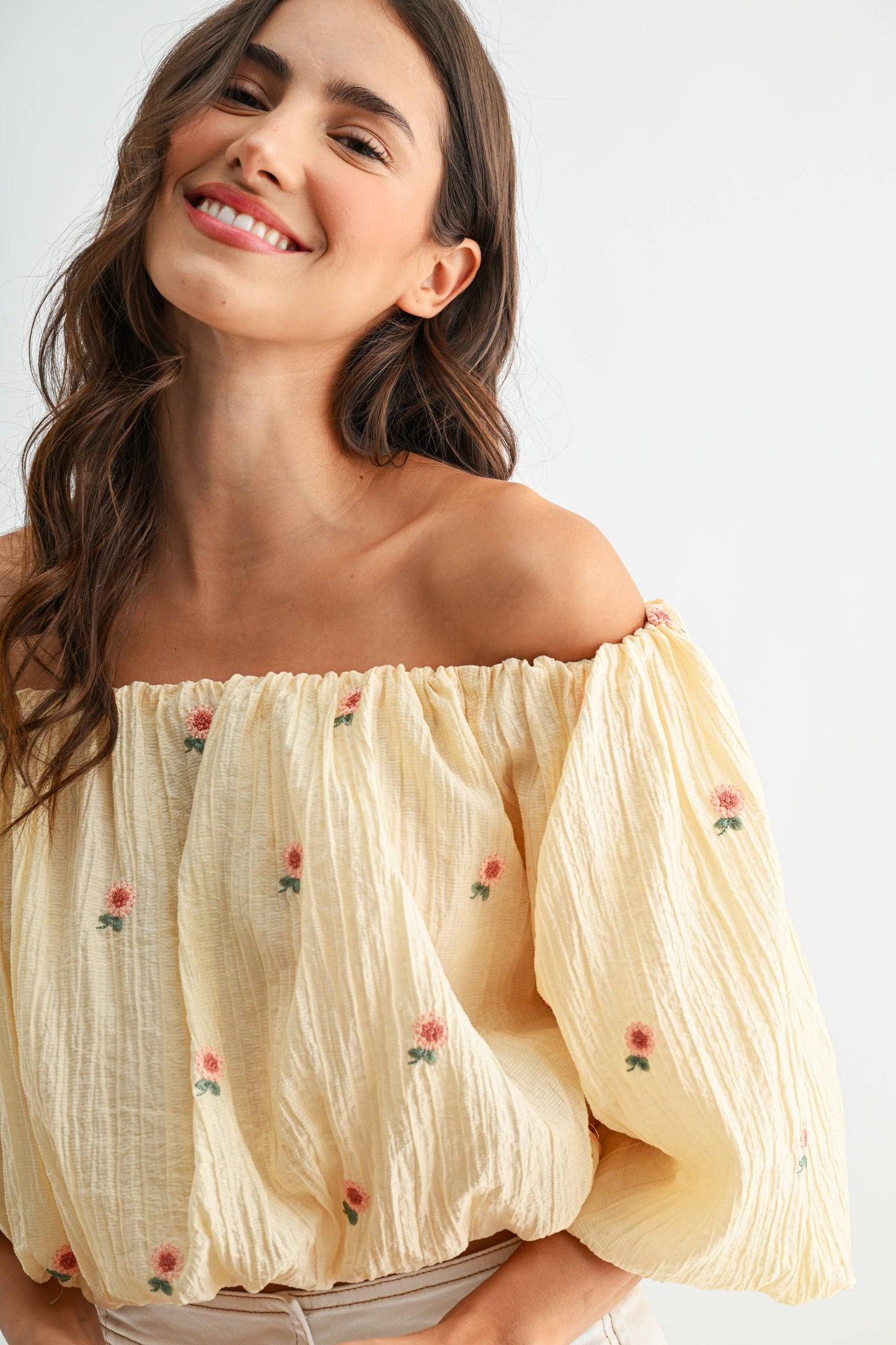 Woman wearing a beige off-shoulder top with floral embroidery on a white background