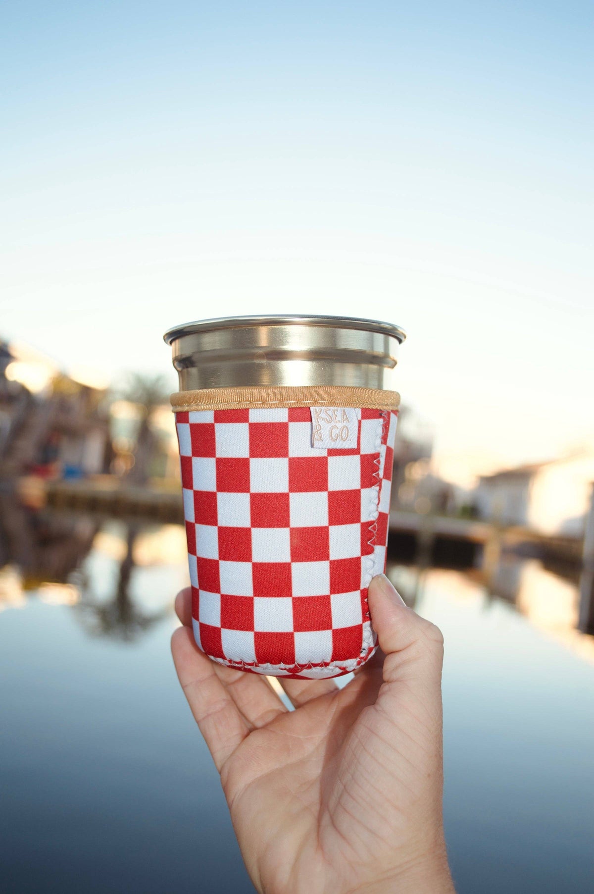 A hand holding a stainless steel travel tumbler with a red and white checkered fabric sleeve, set against a blurred outdoor waterfront background.