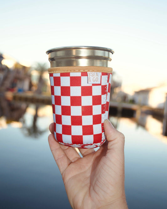 A hand holding a stainless steel travel tumbler with a red and white checkered fabric sleeve, set against a blurred outdoor waterfront background.