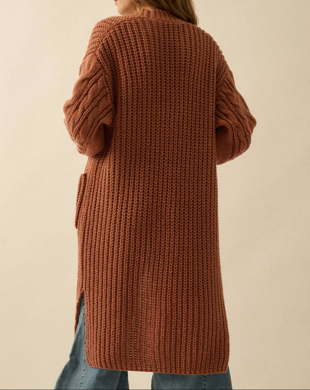 A woman shown from the back wearing a long, chunky ribbed knit cardigan in a warm rust brown color, paired with blue jeans, against a plain beige background.