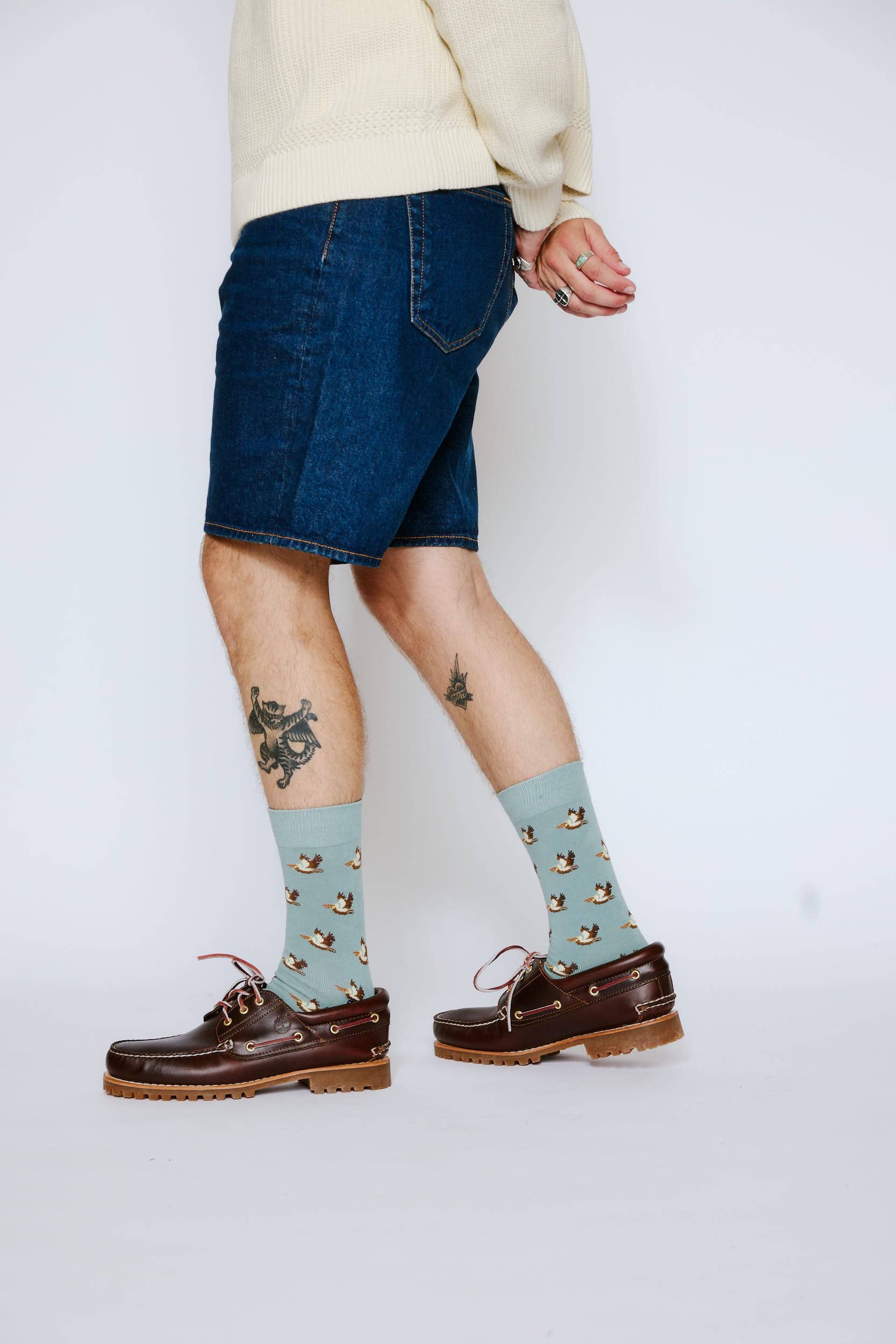 Person wearing denim shorts, cream sweater, brown shoes, and light blue socks with a pattern on a white background