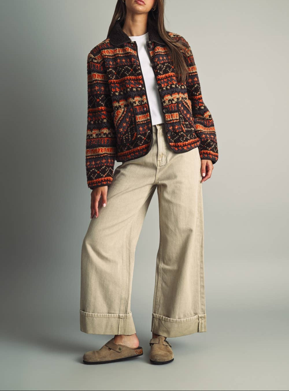 A woman stands against a plain light gray studio background wearing a patterned cropped jacket with orange, black, and red geometric motifs over a white top, wide-leg beige pants, and taupe suede mules.