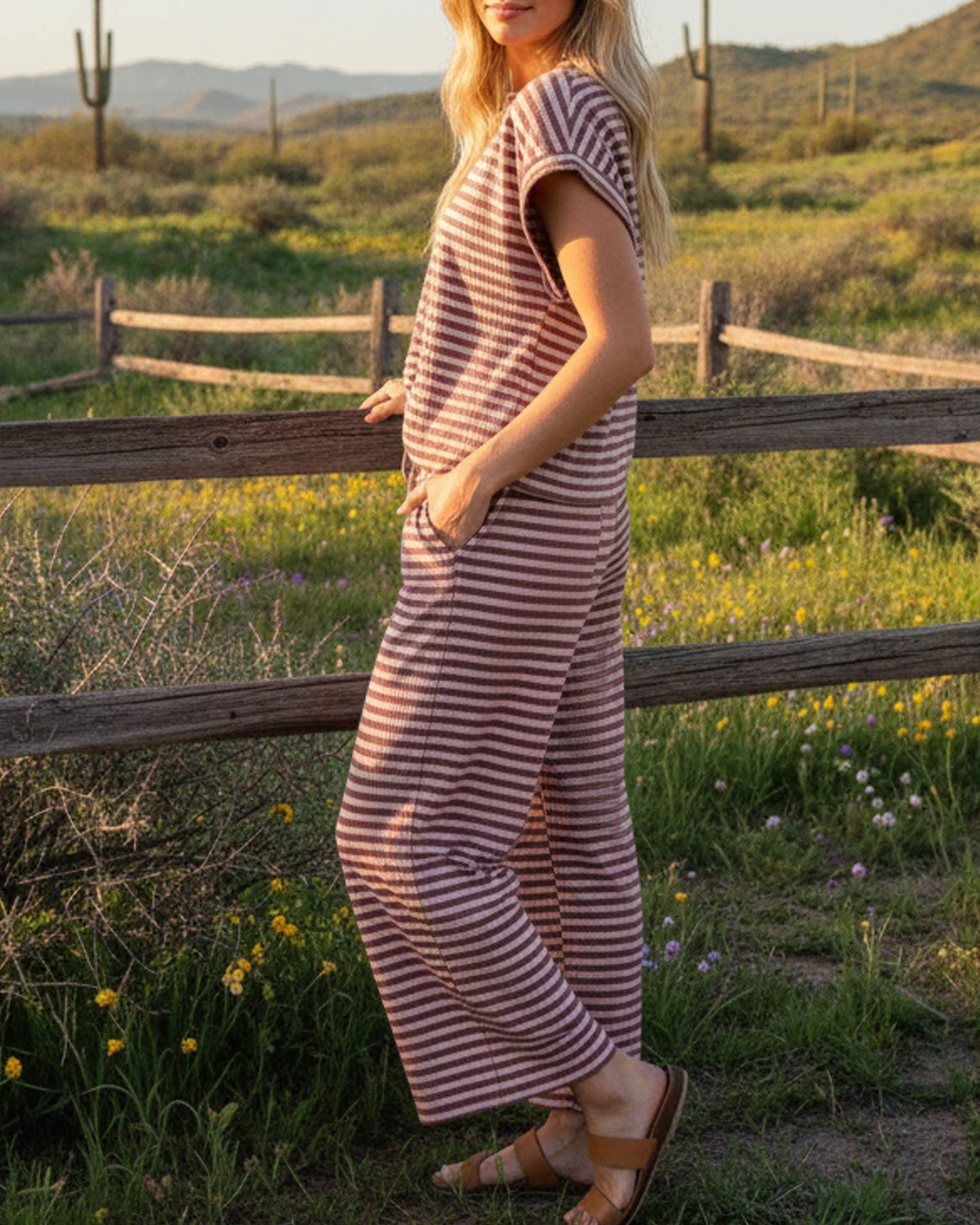 Woman in a striped dress standing in a field with a wooden fence and desert landscape in the background