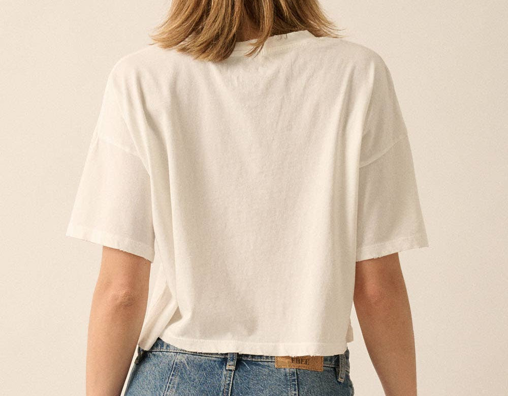 A woman viewed from behind wearing an oversized off-white short-sleeve T-shirt and blue jeans against a plain beige studio background.