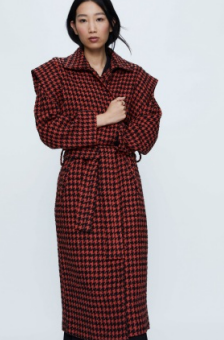 A model wearing a long checkered coat with shoulder pads in black and coral, with a belt tied at the waist.