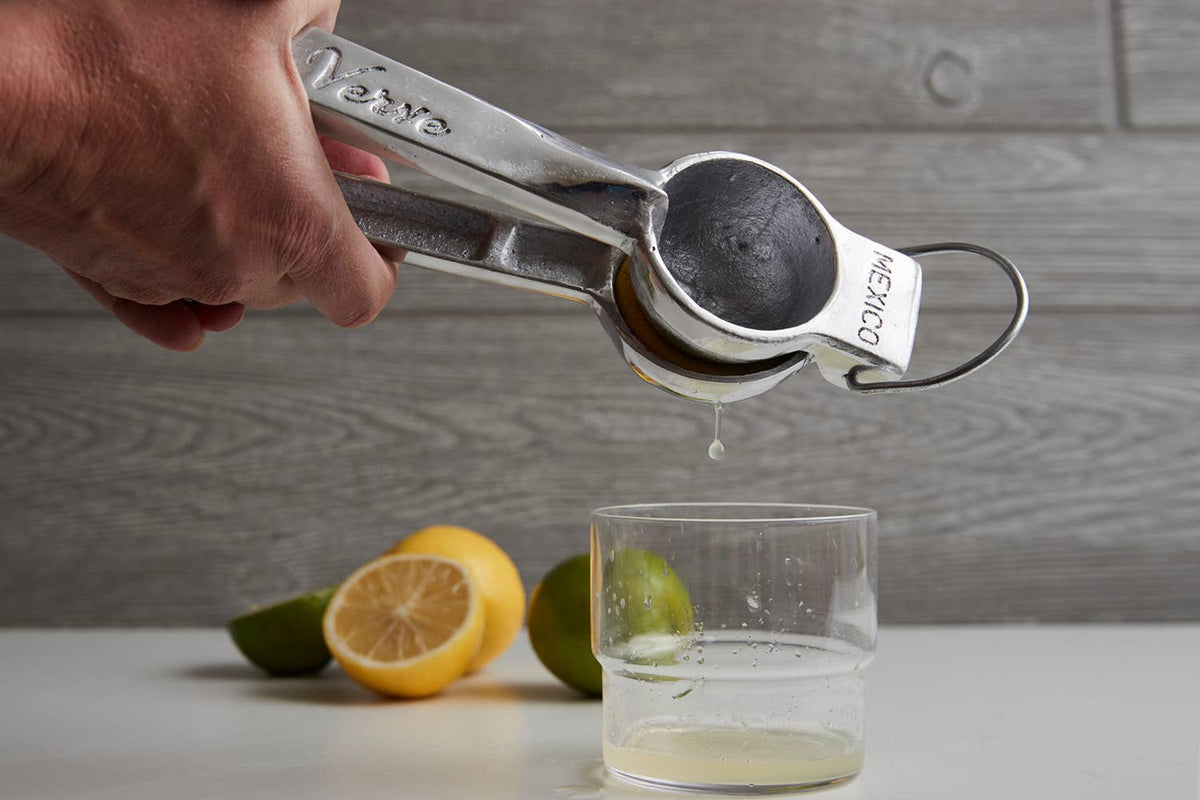 Hand using a citrus press to extract juice from a lime into a glass with lemons and limes on a wooden surface.