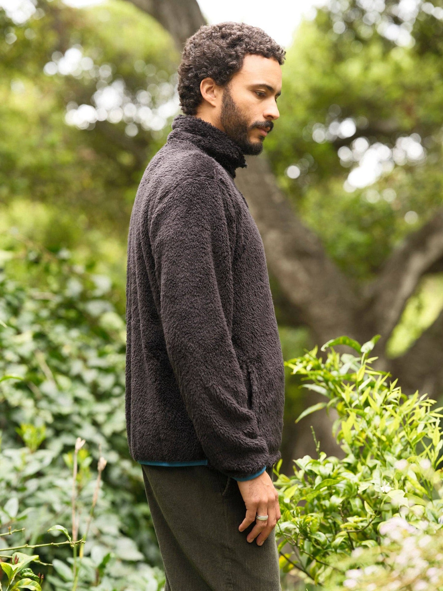 Man wearing a dark sweater standing in a natural setting with trees and greenery.