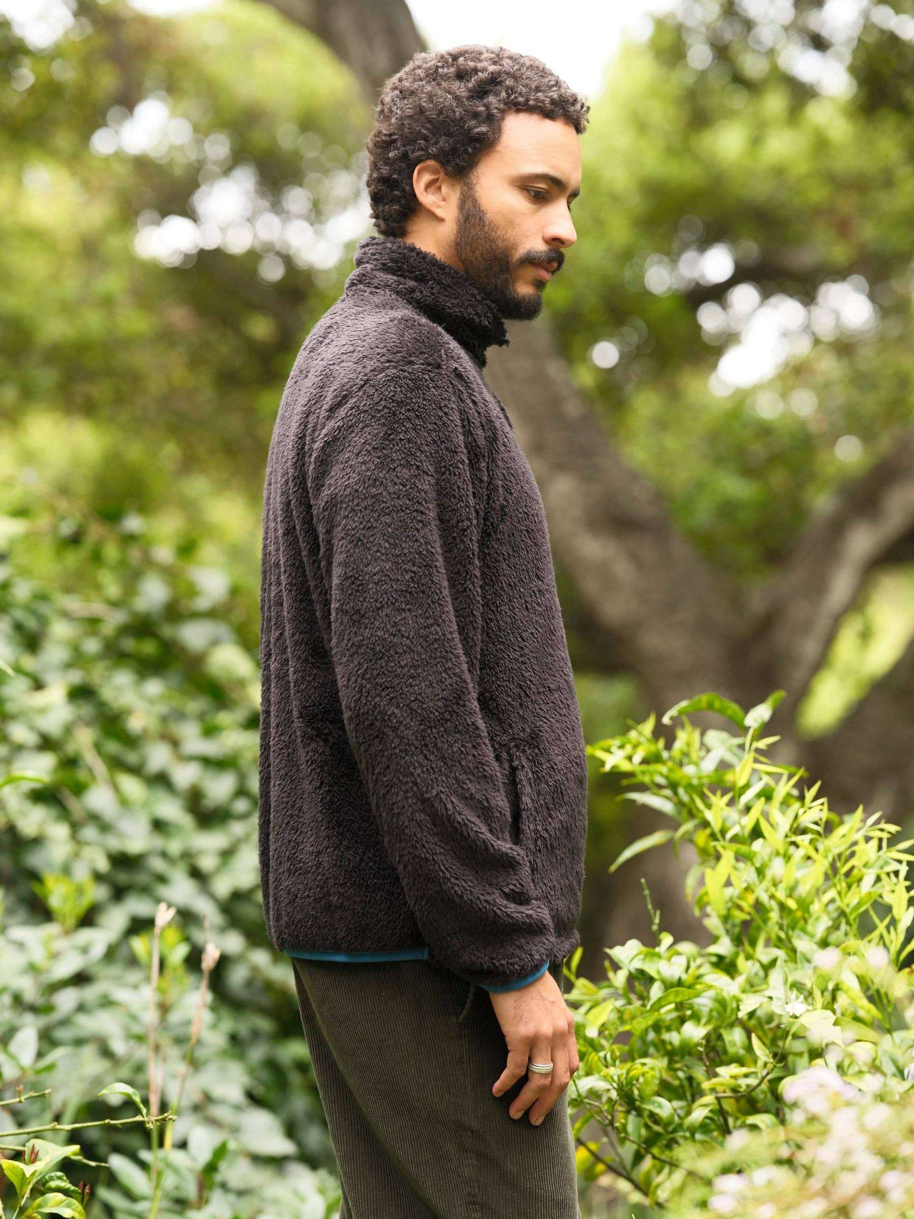 Man wearing a dark sweater standing in a natural setting with trees and greenery.