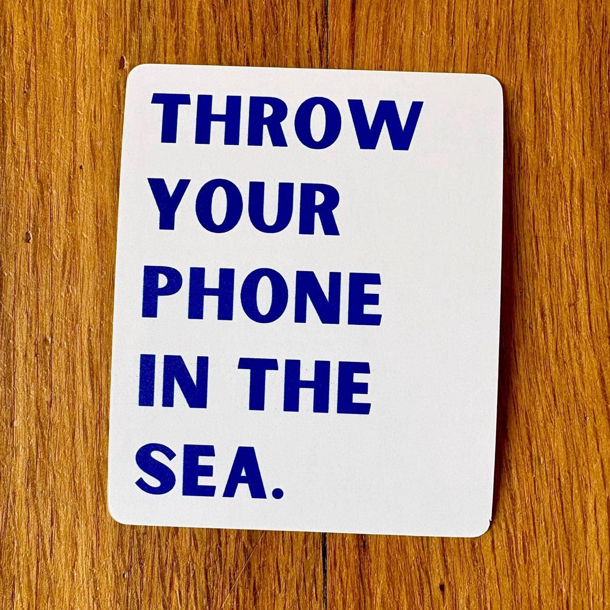 White card with blue text 'THROW YOUR PHONE IN THE SEA' on a wooden surface
