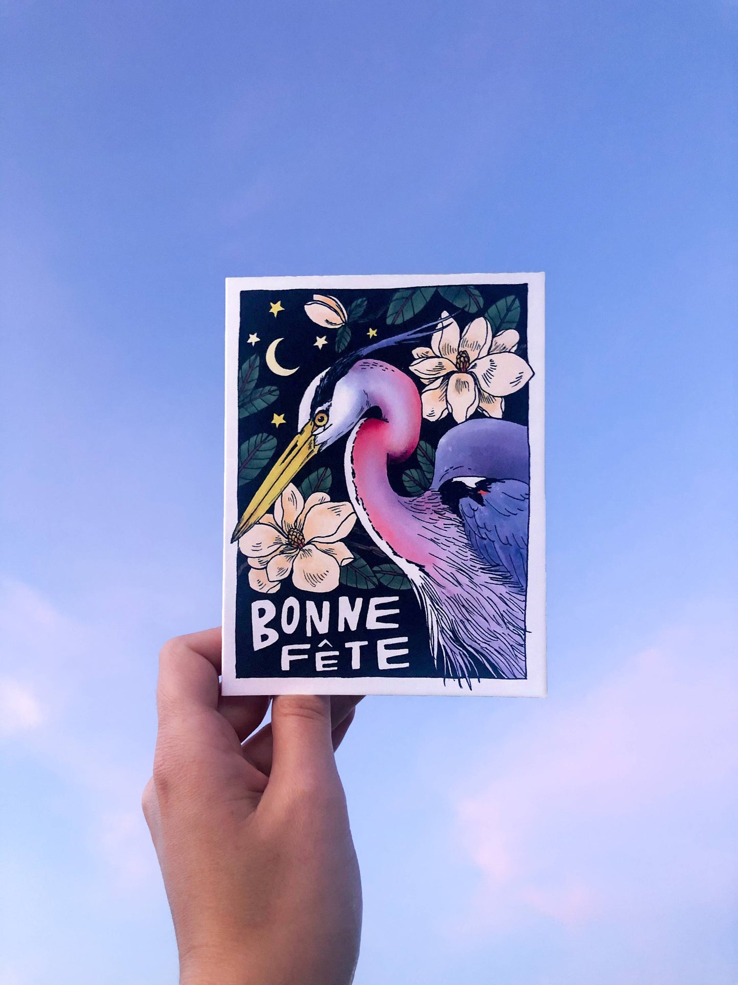 Hand holding a card with a heron illustration and 'Bonne Fète' text against a blue sky.