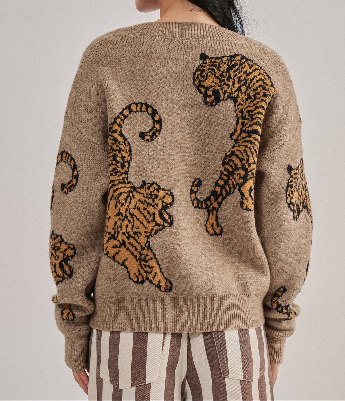 A woman shown from the back wearing a beige knit sweater with large tiger motifs in tan and black, paired with brown and white striped pants, against a light gray studio background.