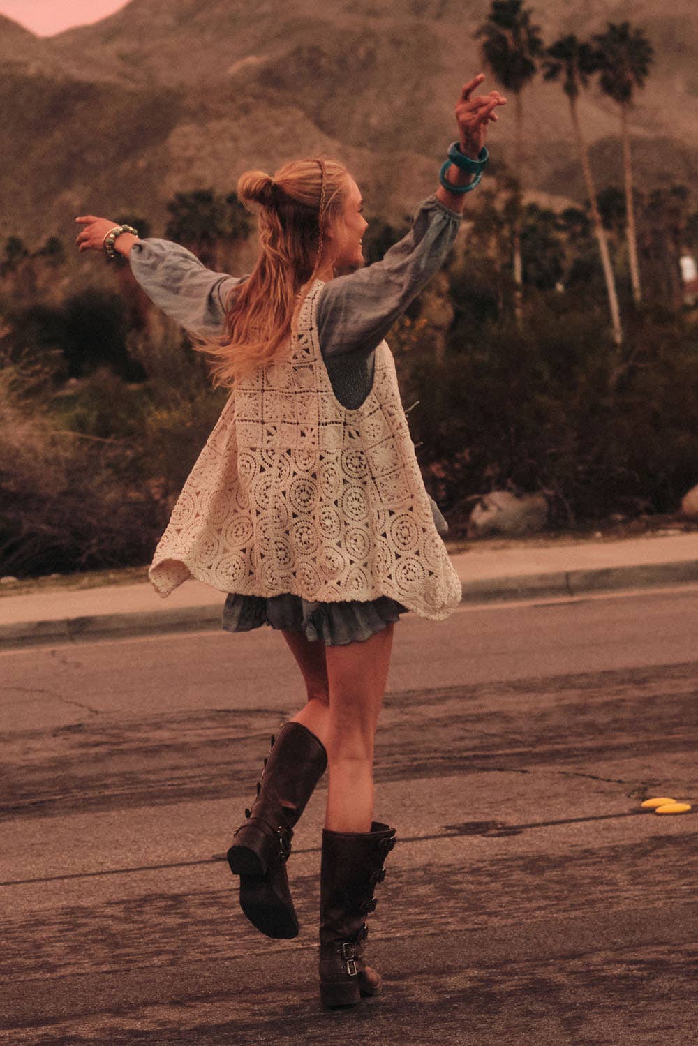 Woman in a lace dress and boots standing on a street with mountains and palm trees in the background