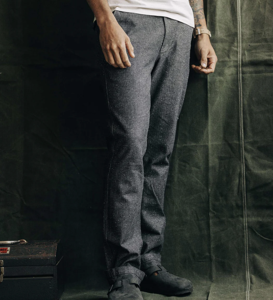 A person standing side-on, wearing The Carnegie Pant in granite herringbone pattern, paired with a white shirt and black shoes, against a green backdrop.