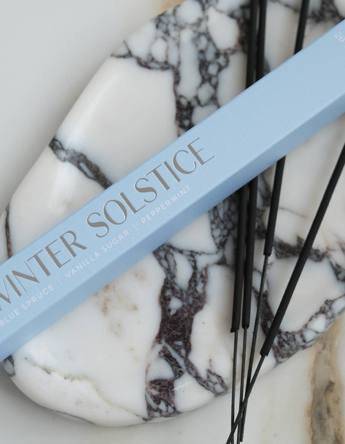 A close-up of a light blue rectangular product box labeled 'WINTER SOLSTICE' resting on a white and gray marble stone, with several black thin sticks placed beside it.