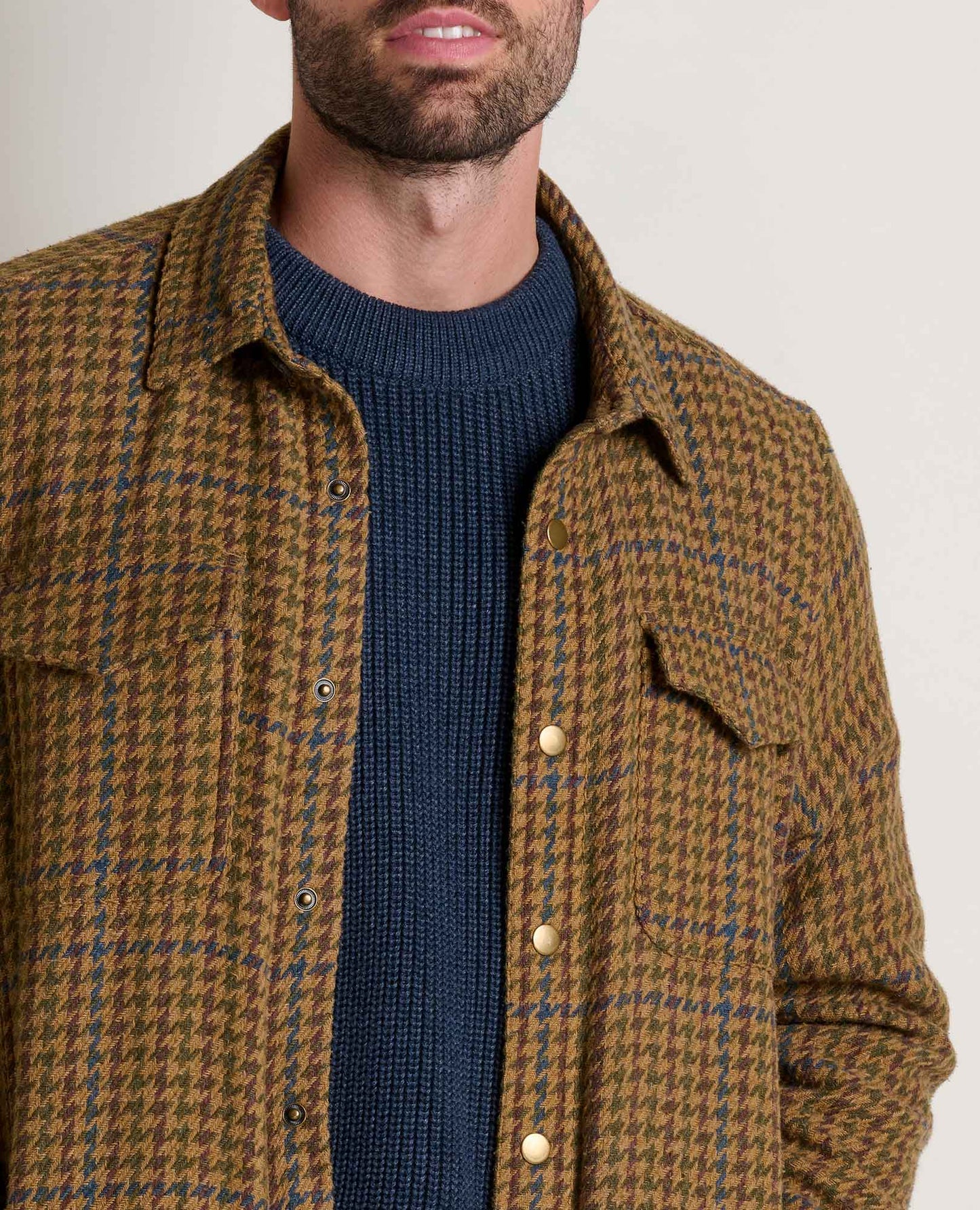 Man wearing a brown checkered shirt over a blue sweater on a plain background