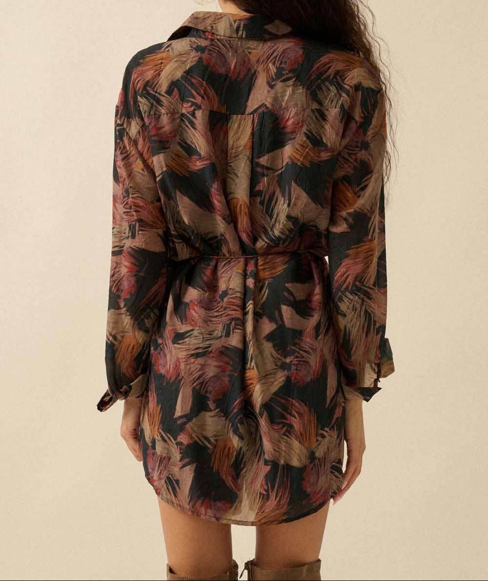 A woman viewed from behind wearing a long-sleeve collared shirt dress with a tropical leaf print in earthy tones of brown, beige, and black, set against a plain beige background.