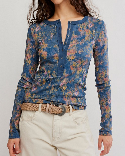 A woman wearing a long-sleeve floral henley top in blue with pink, yellow, and green floral print, paired with cream pants and a woven belt.