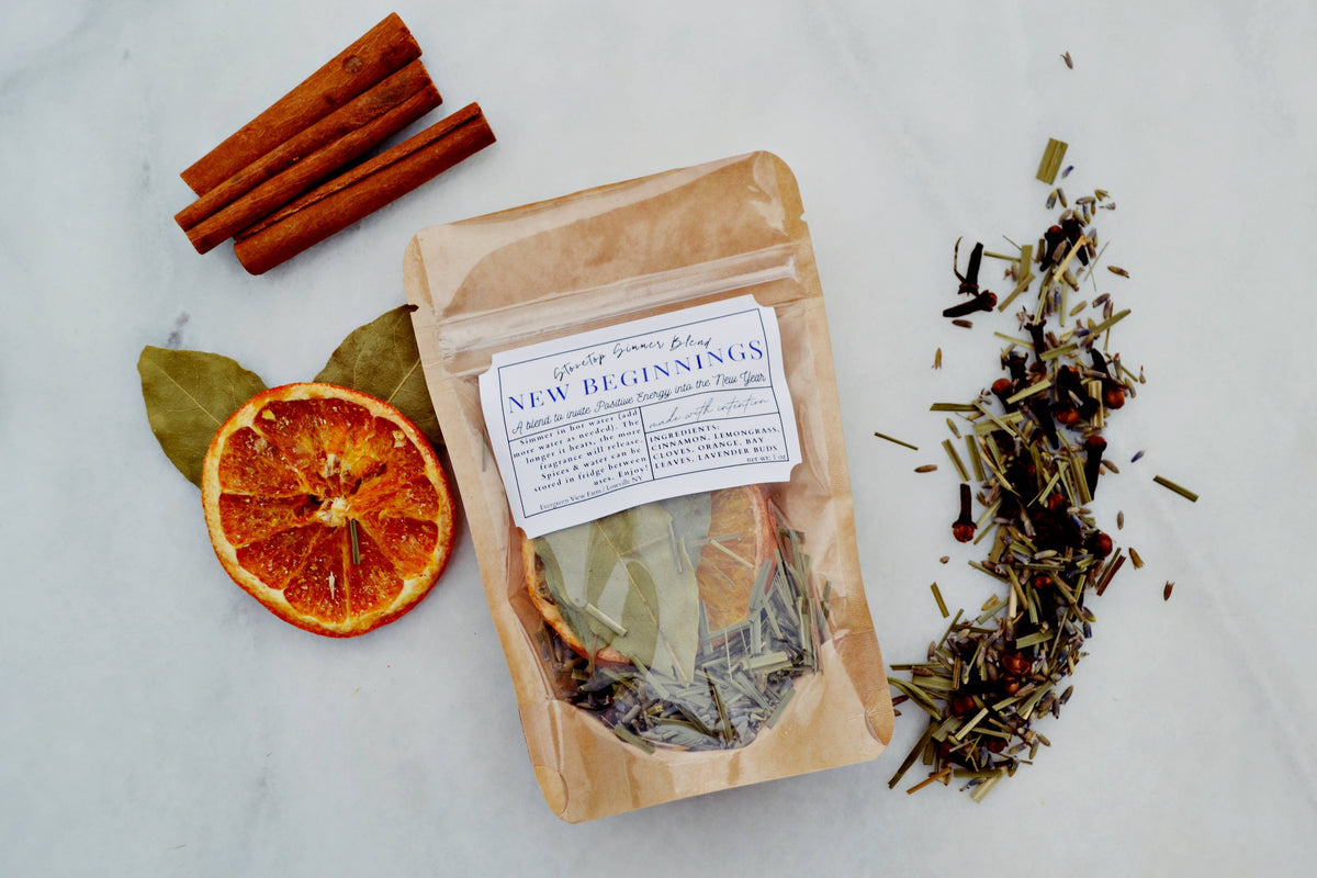 A kraft paper pouch labeled 'New Beginnings' herbal tea blend sits on a light marble surface, surrounded by cinnamon sticks, a dried orange slice, bay leaves, and loose dried herbs.