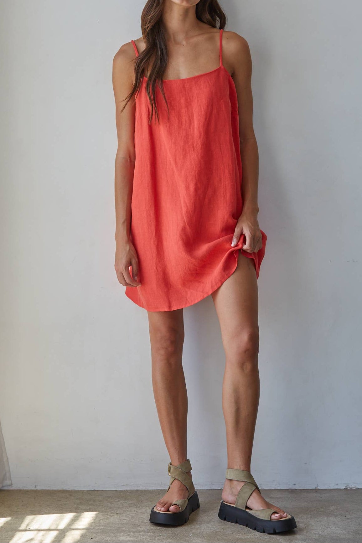 A woman modeling a loose, coral-red linen slip dress with thin straps and black platform sandals, standing against a plain white wall.