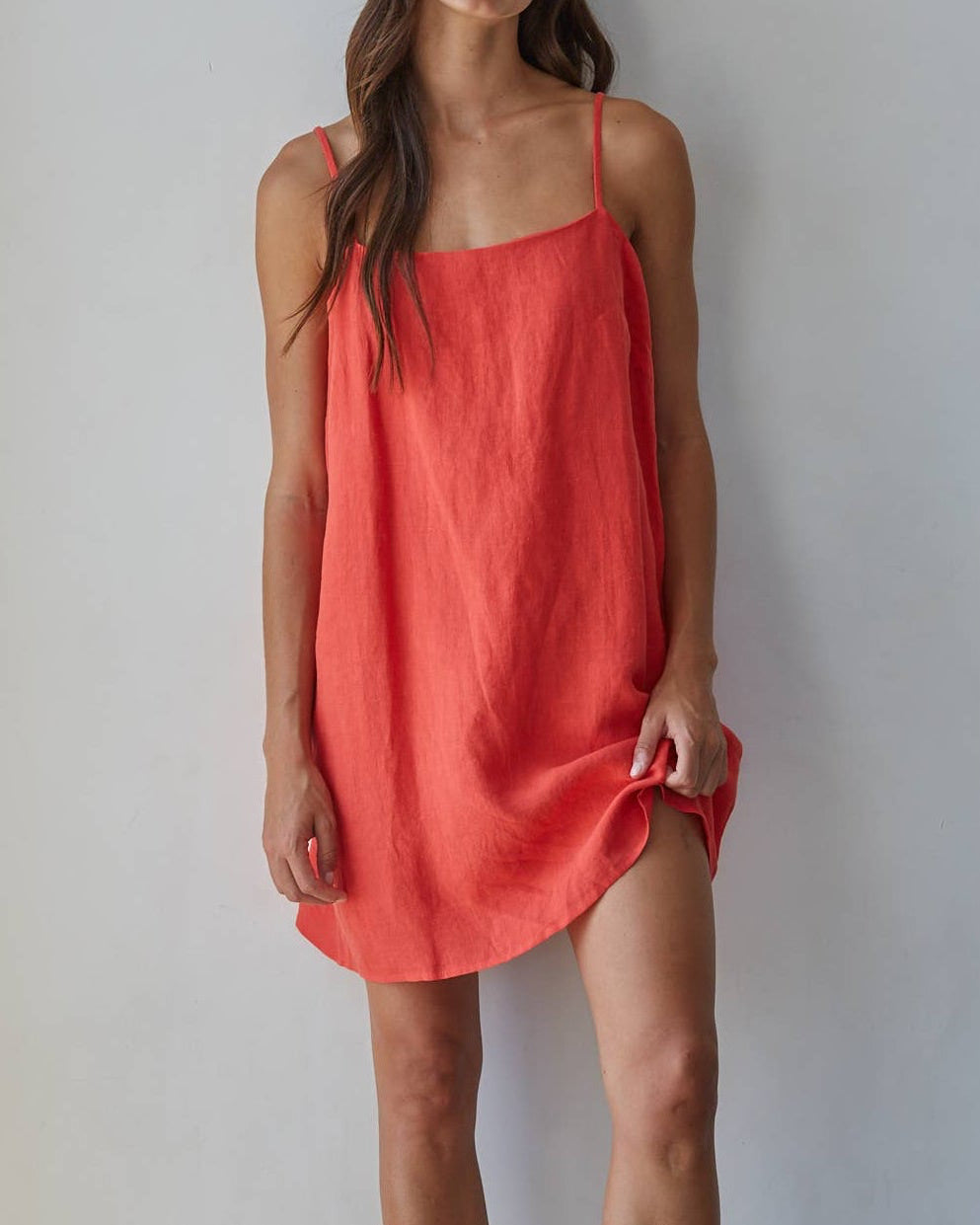 A woman modeling a loose, coral-red linen slip dress with thin straps and black platform sandals, standing against a plain white wall.