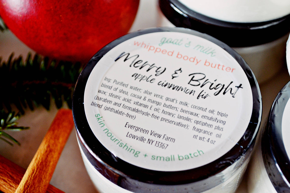 A close-up of a jar of whipped body butter labeled 'Merry & Bright apple cinnamon pie' by Evergreen View Farm, with a cinnamon stick and red apple props in a styled setting.