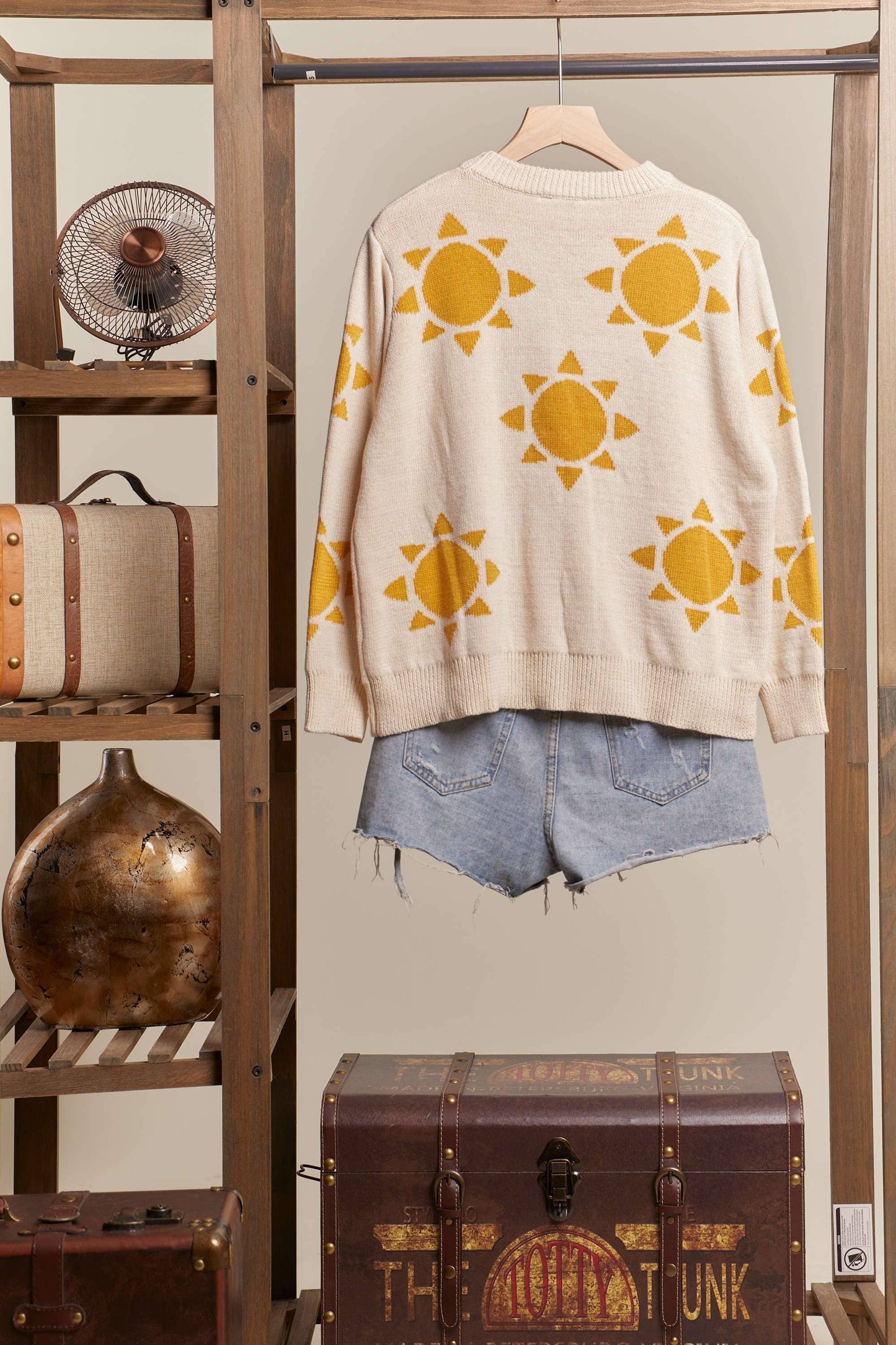 Beige sweater with yellow sun patterns hanging on a rack with denim shorts below, against a neutral wall.
