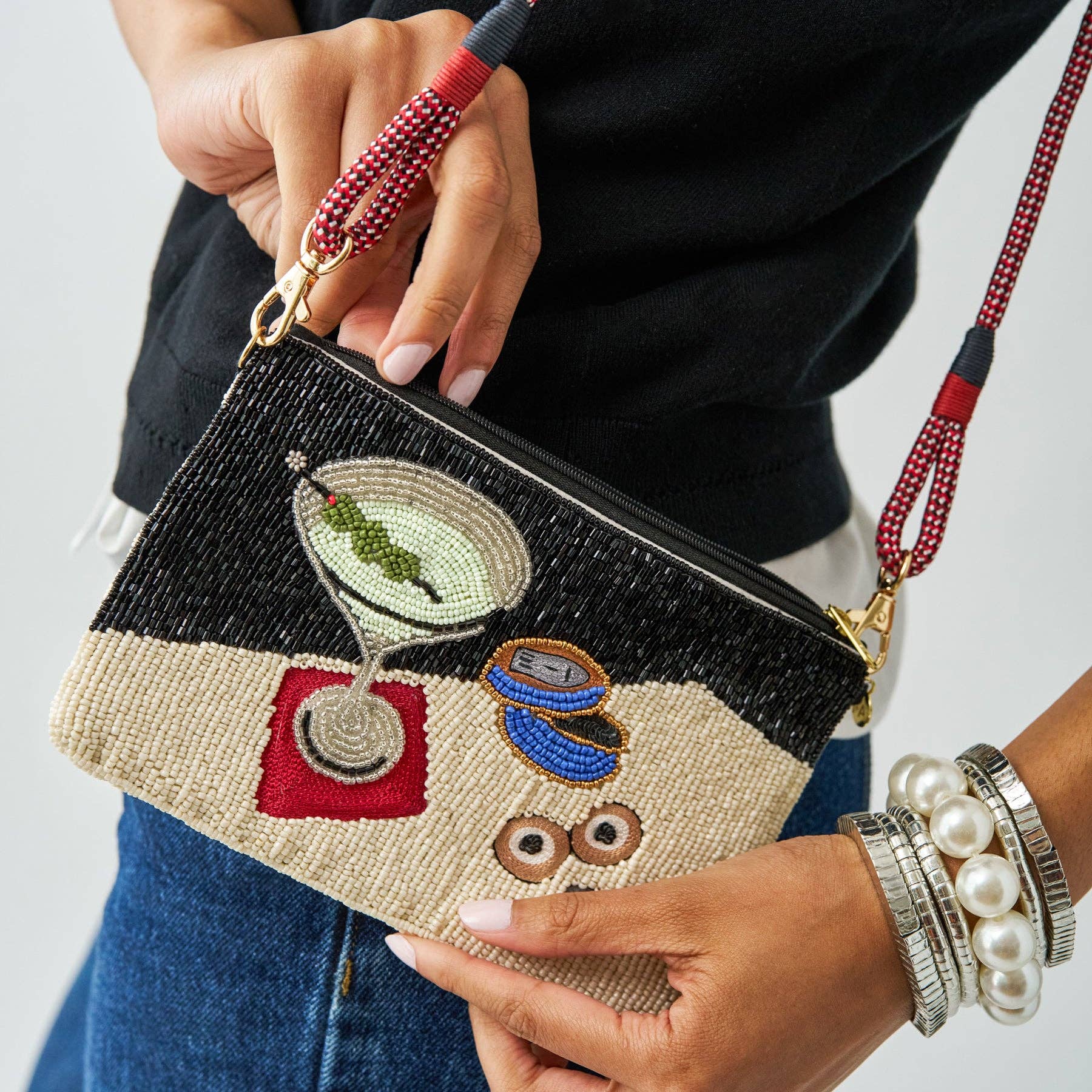 A close-up of a person holding a small rectangular clutch bag with a black and beige woven body, embroidered cocktail motifs, gold-tone hardware, and a red-and-black beaded wrist strap.