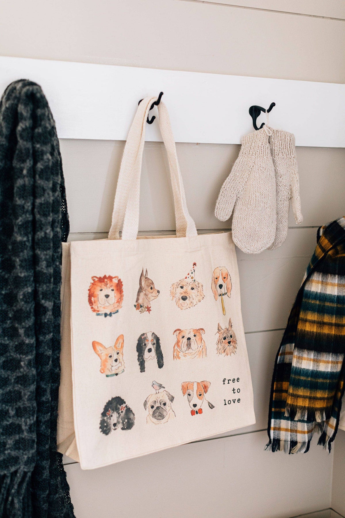 A cream canvas tote bag with watercolor dog illustrations hanging on a white wall hook, next to a gray patterned blanket, knit mittens, and a plaid scarf.
