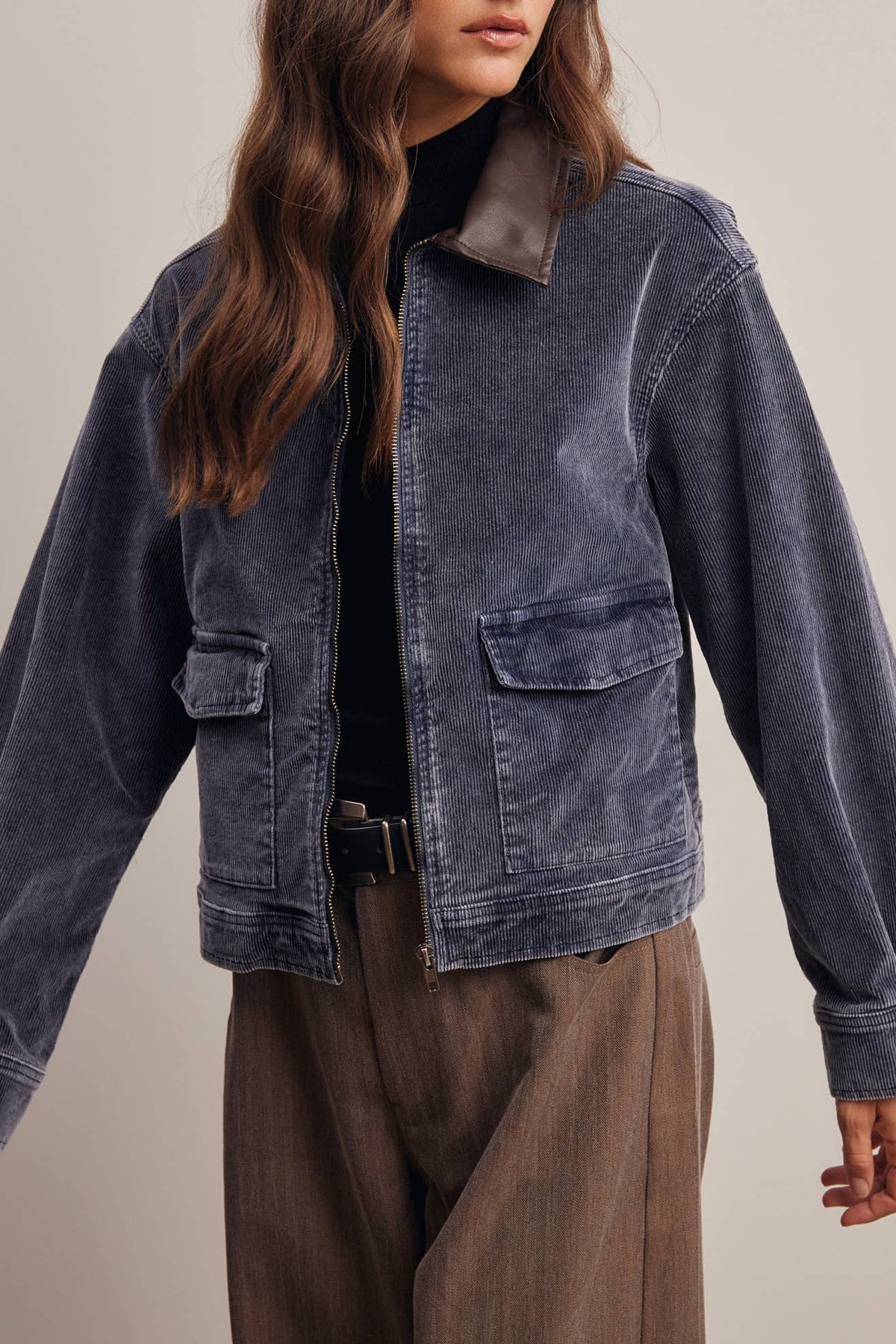 A woman wearing a dark blue corduroy jacket with a brown collar, styled with a black top and brown trousers, photographed against a neutral beige background.