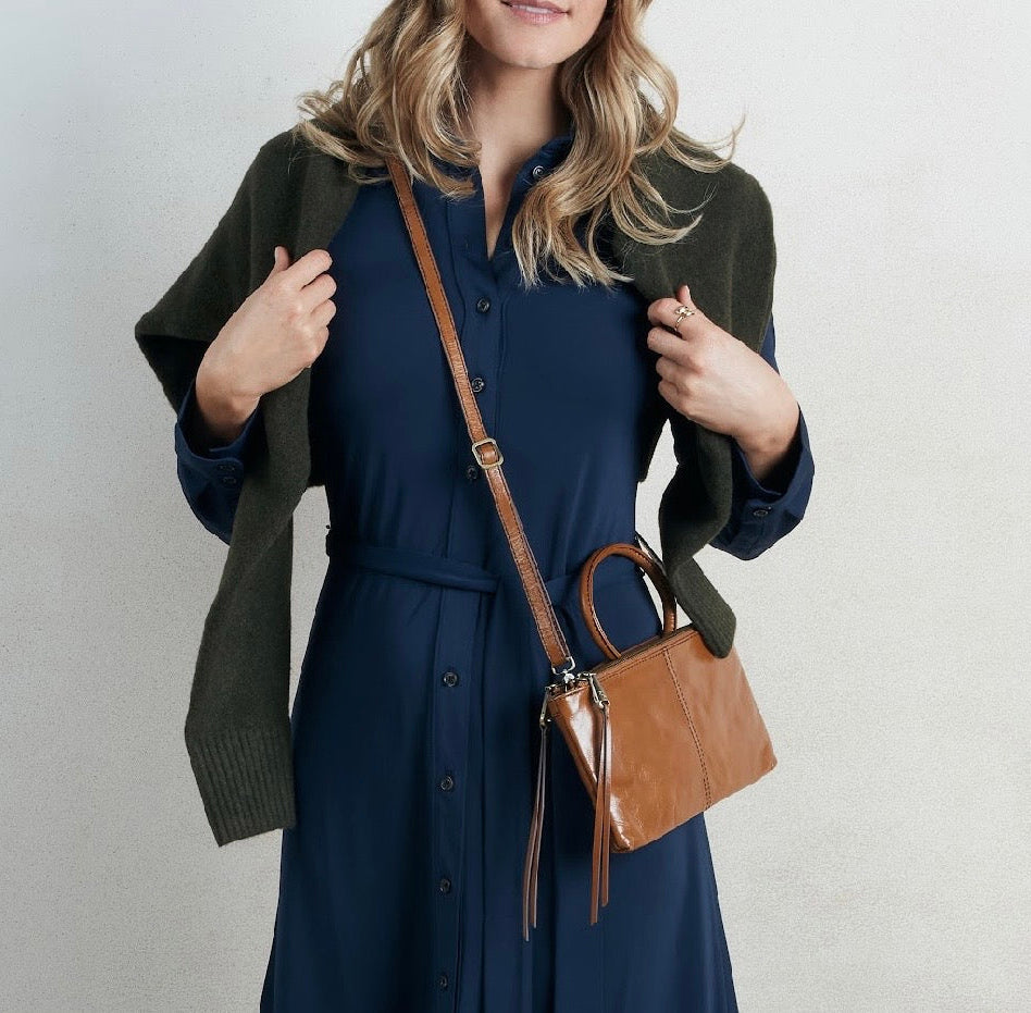 A woman wearing a navy blue dress, an olive green cardigan, and a brown leather crossbody handbag, standing against a light gray wall.