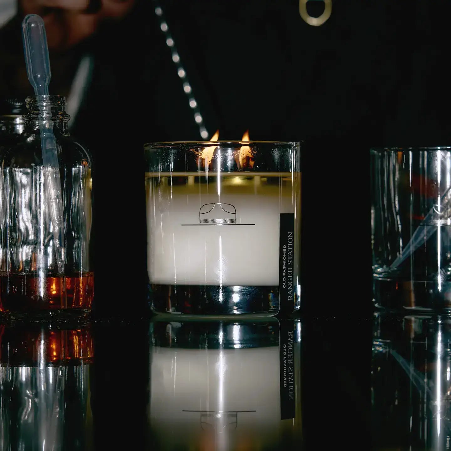 Candle in a glass container on a reflective surface with bottles and glasses in the background.