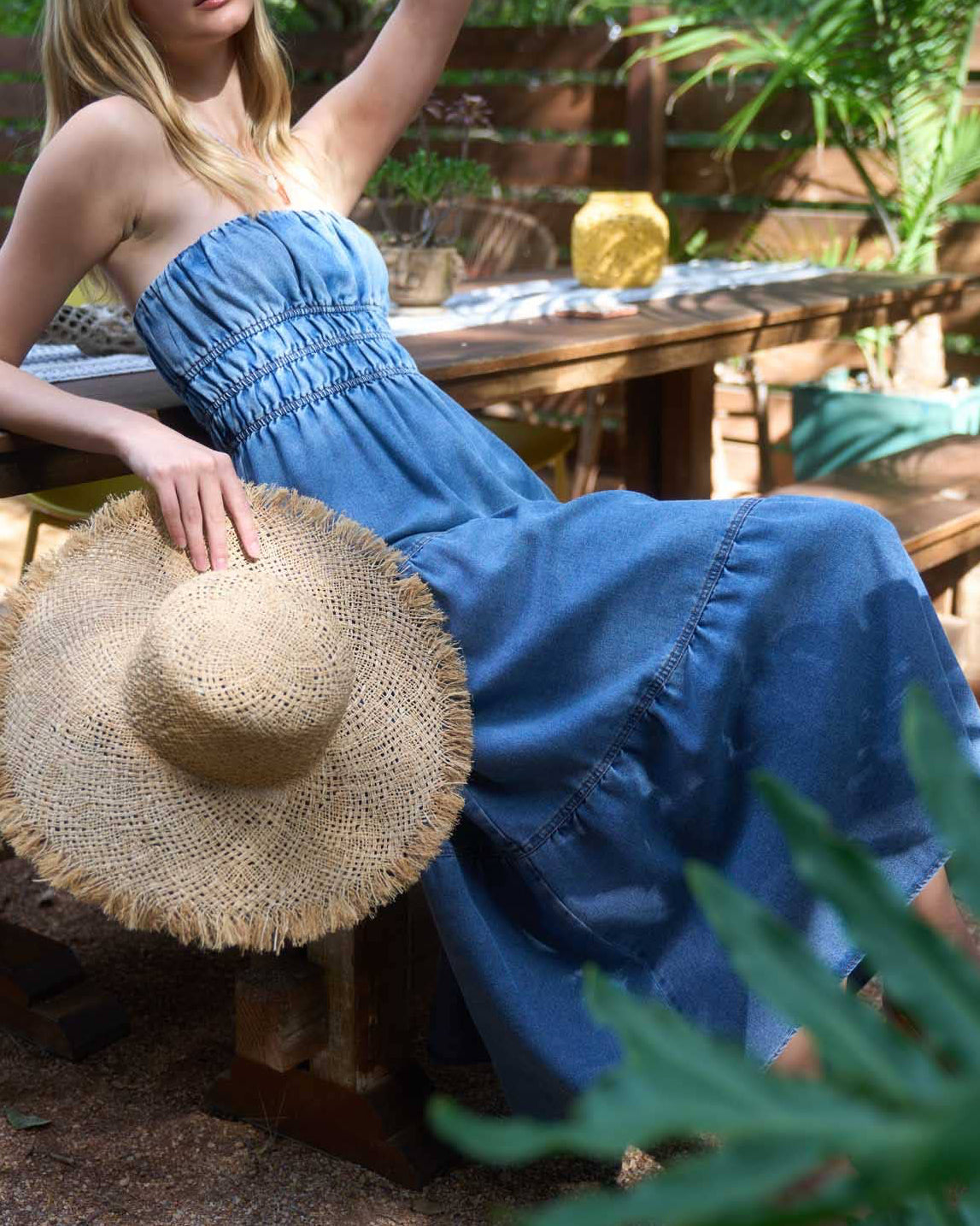 Woman in a blue dress sitting outdoors with a straw hat on a wooden table.