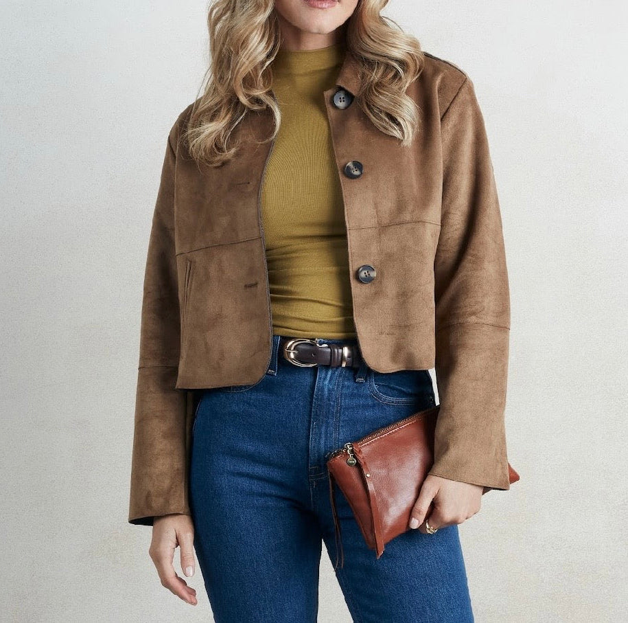 A woman wearing a cropped brown suede jacket over a mustard yellow ribbed top, paired with blue jeans and holding a brown leather clutch against a light gray background.
